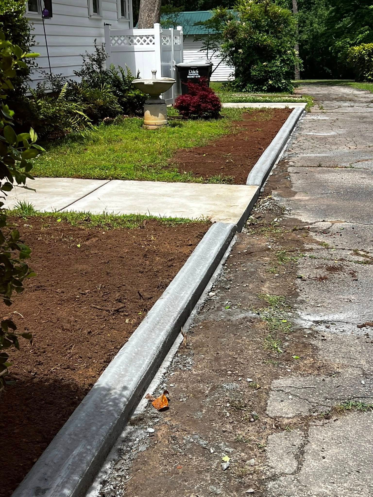 Concrete curb separates a driveway from a flower bed and walkway, with a house in the background.
