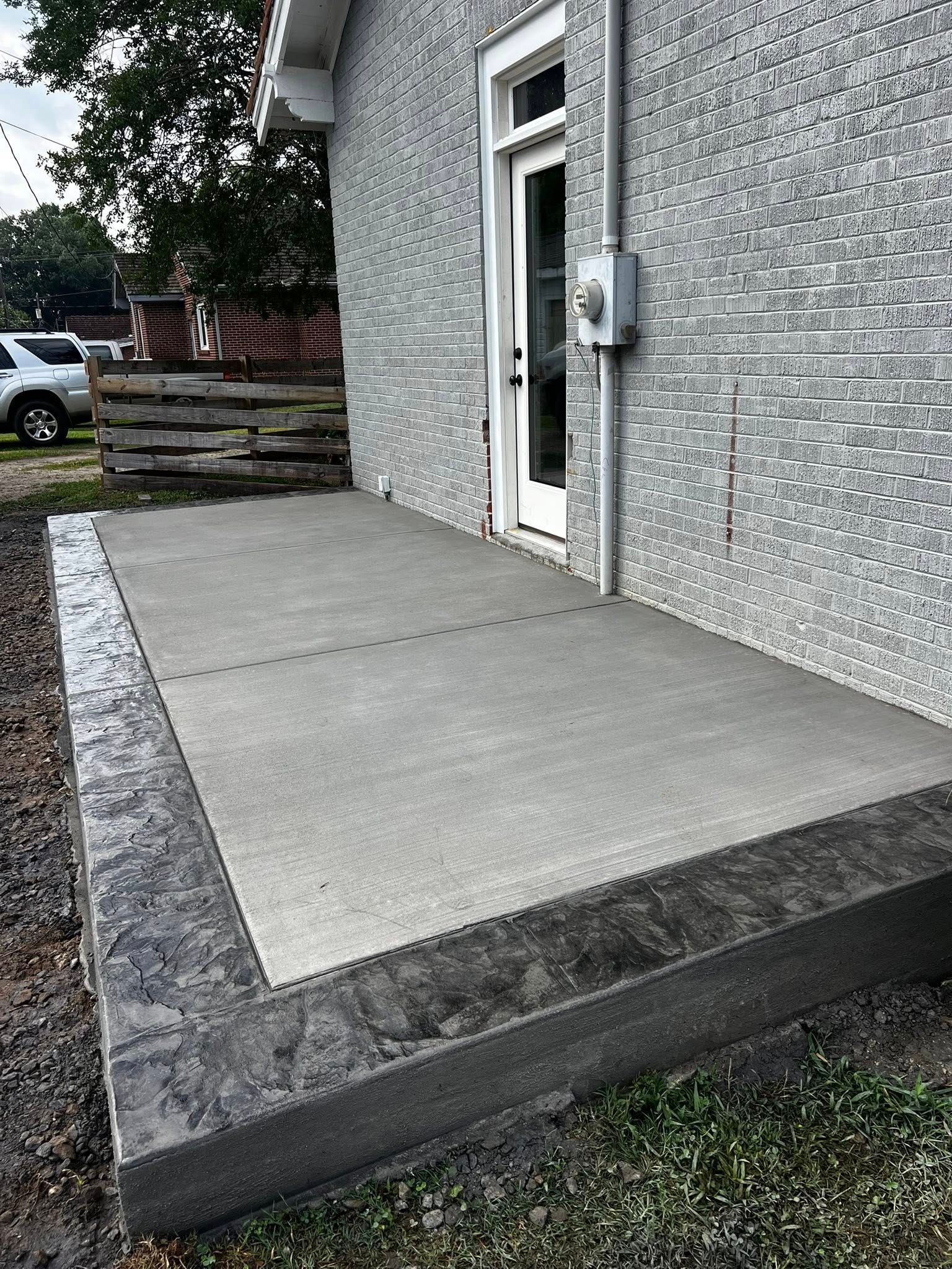 Concrete patio with a darker stamped border, next to a brick house, near a door.