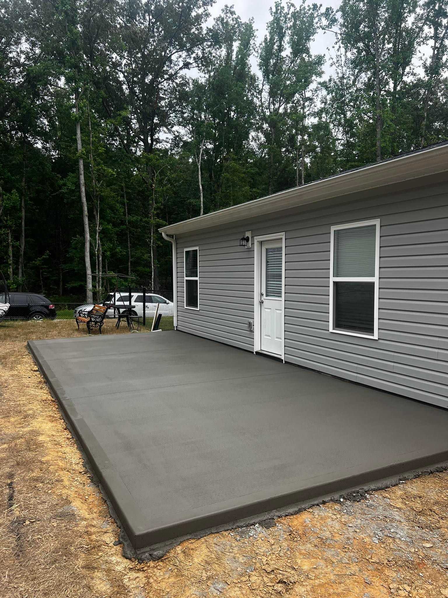 A concrete patio next to a gray house with a white door, surrounded by dirt and trees.
