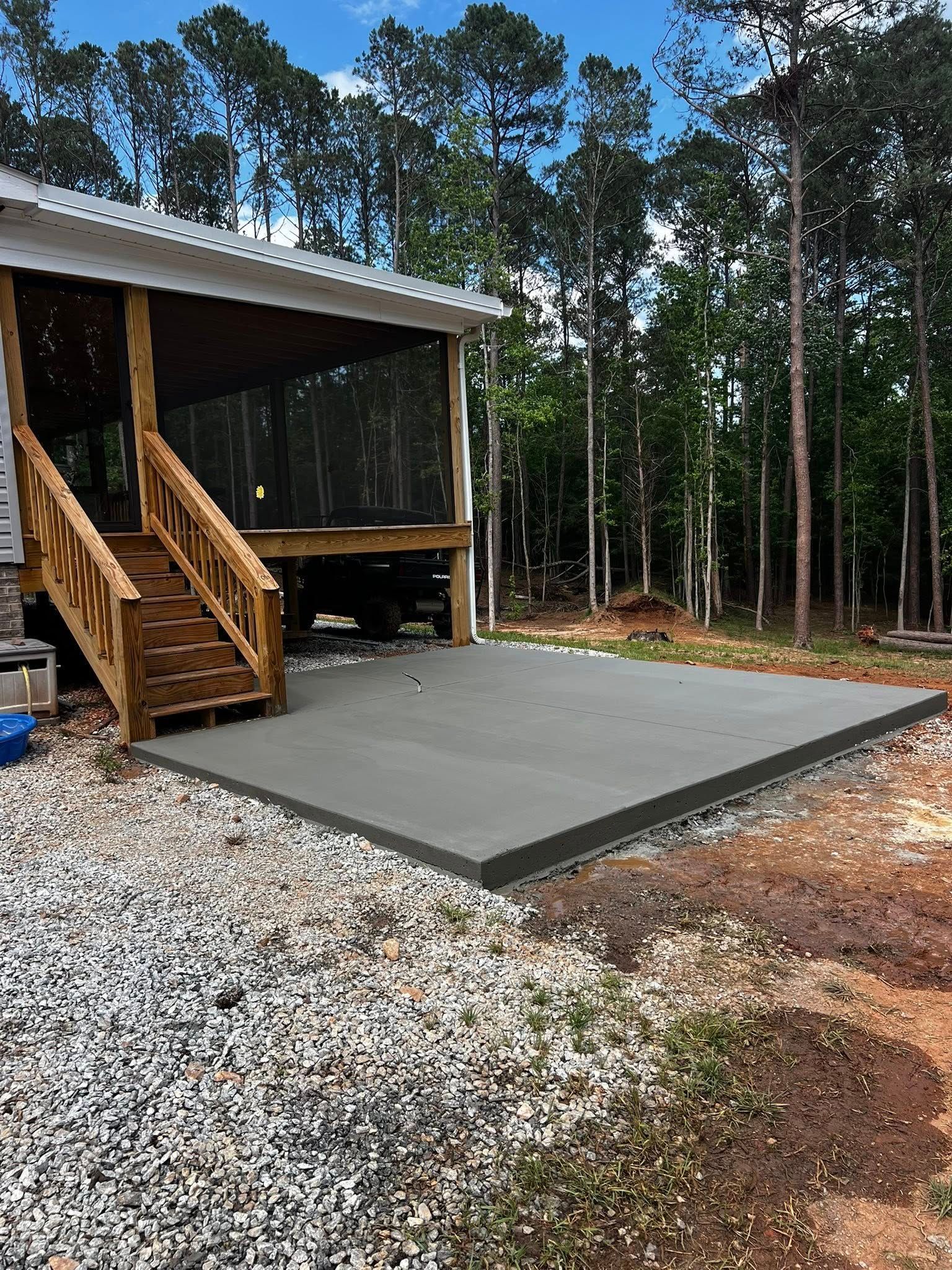 Newly poured concrete patio next to a screened porch with wooden stairs, in a wooded area.