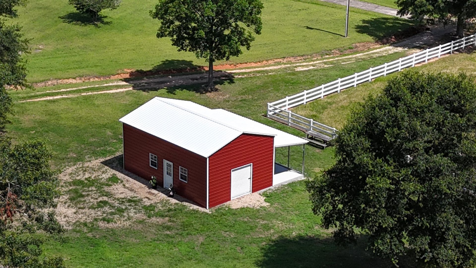 Red barn with white roof and awning on green field, white fence in background.