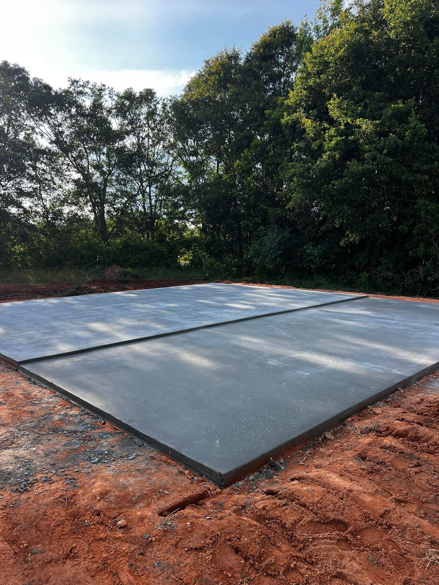 Freshly poured concrete slab, set in a red clay yard, with trees in the background.