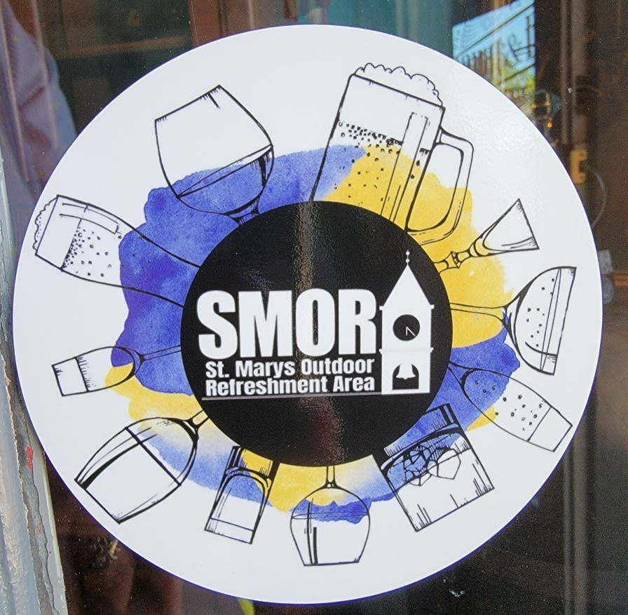 Circular sign for SMOR (St. Mary's Outdoor Refreshment Area) with glasses and beer mugs .