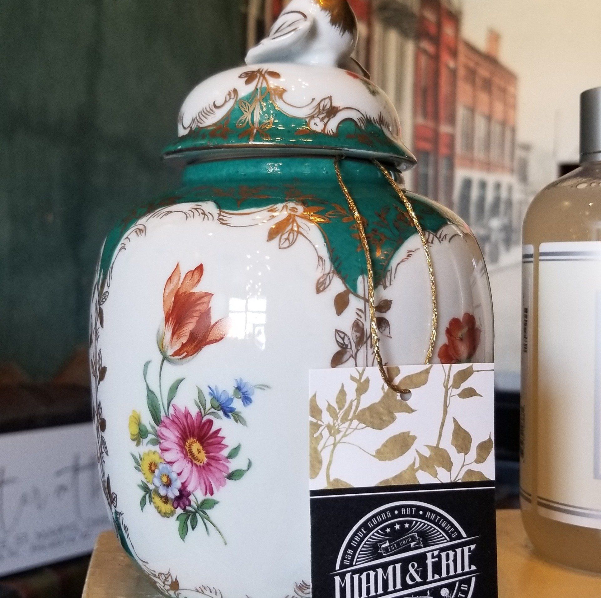 A floral-decorated ceramic jar with a bird finial and a gold necklace, on a shelf next to a label and a bottle.