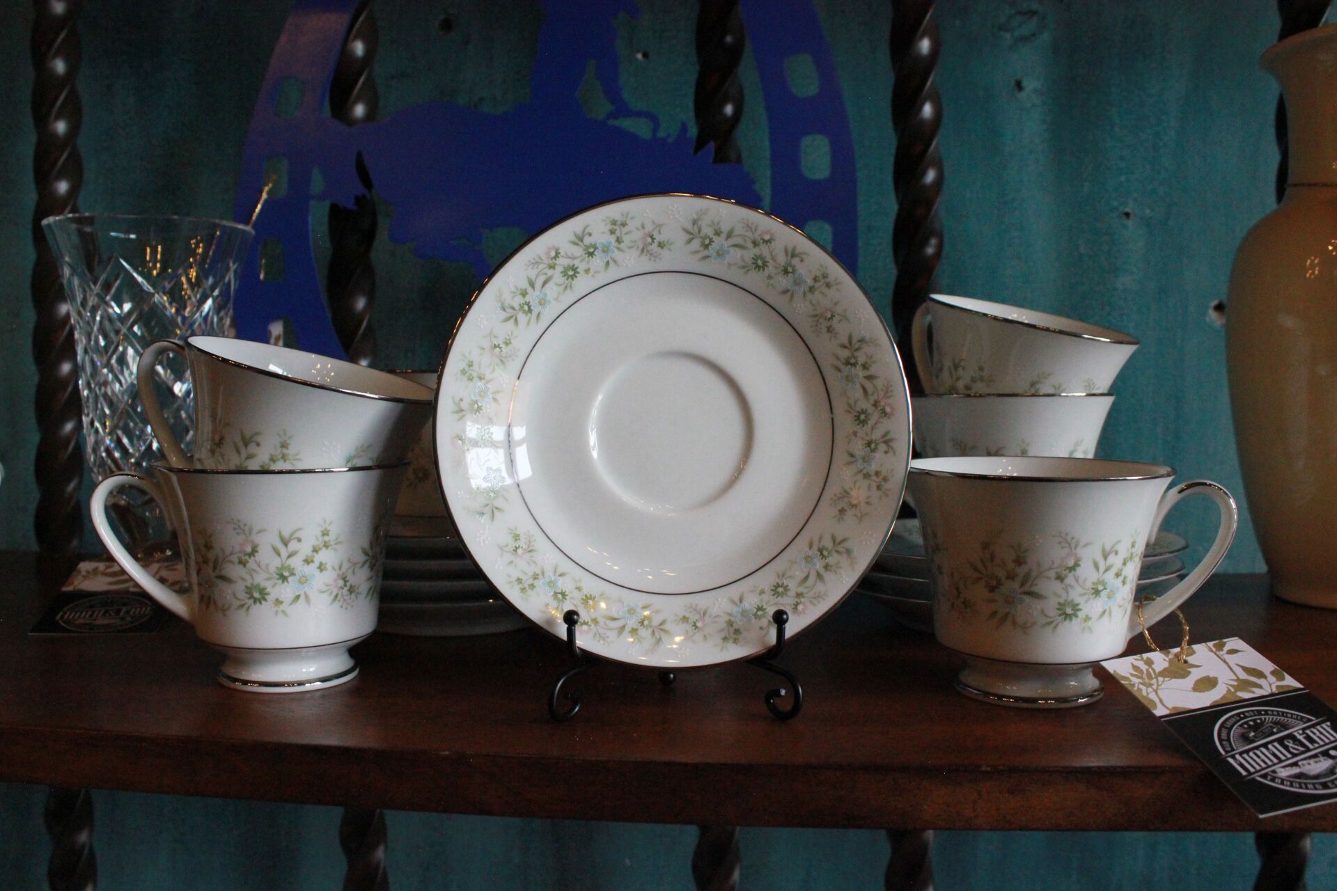 White teacups and saucers with floral design sit on a wooden shelf.
