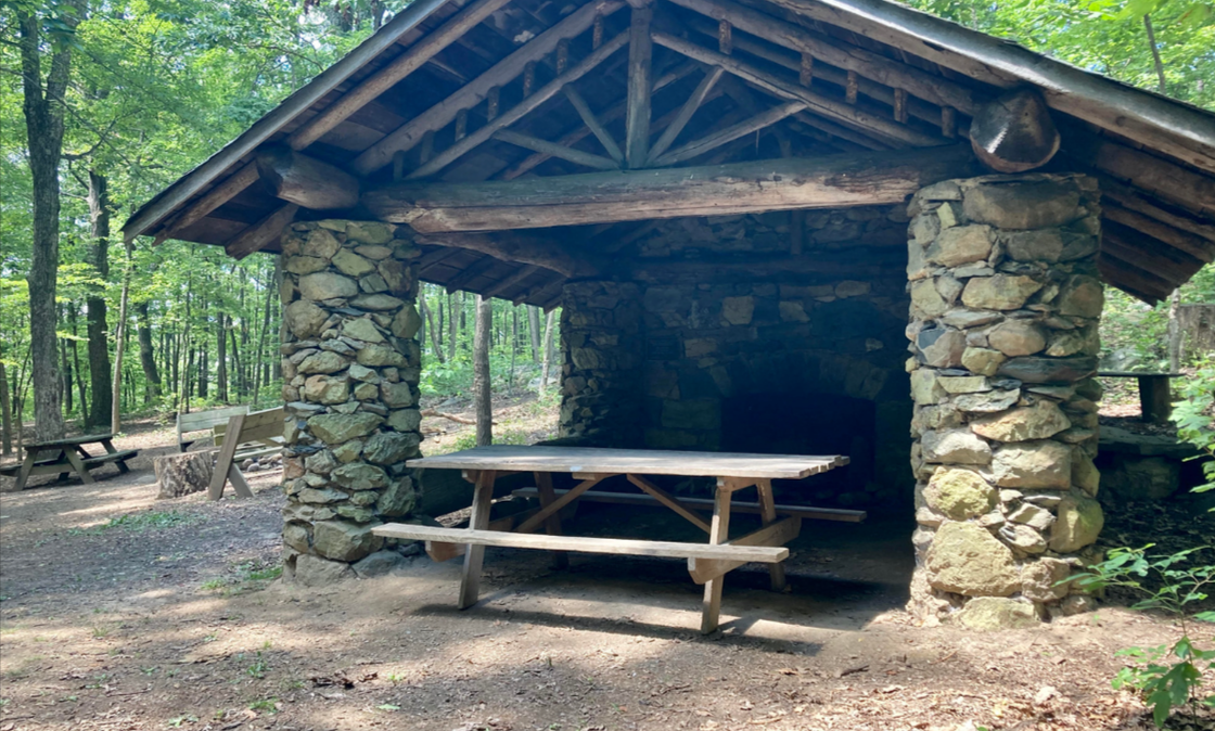A stone shelter with a picnic table inside of it in the woods.