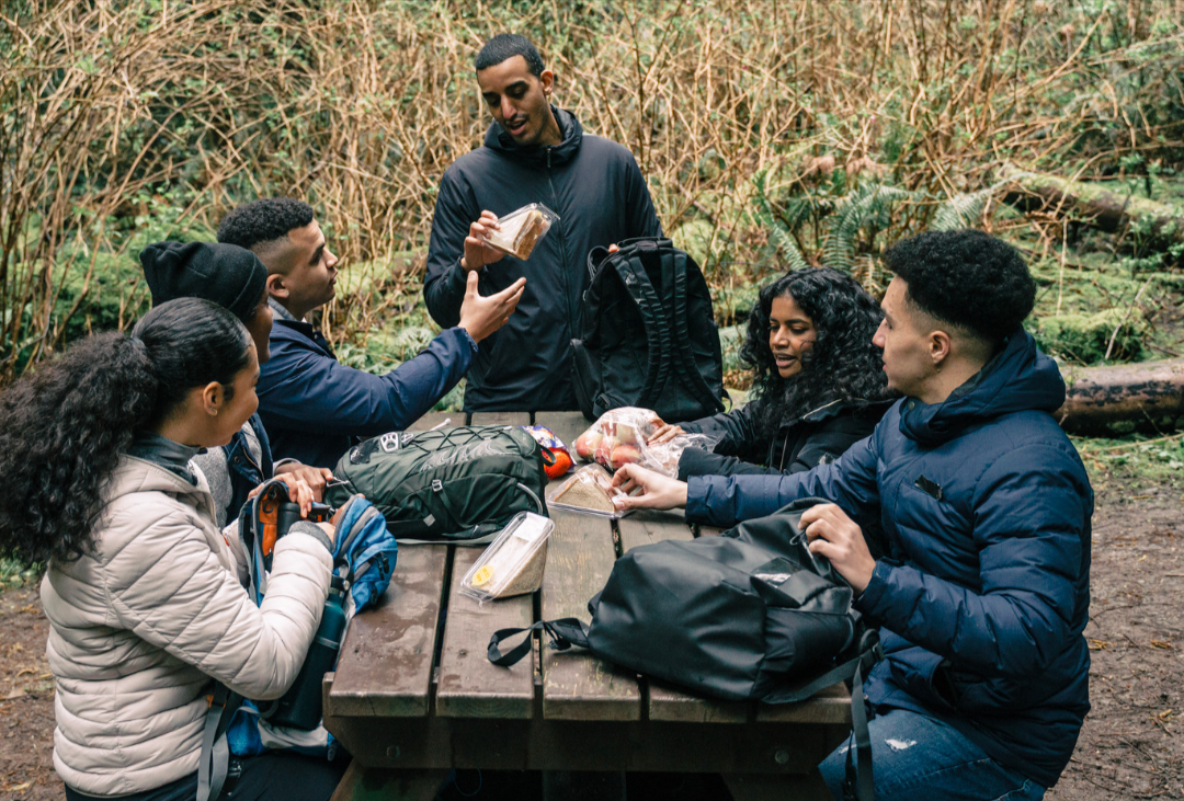 A group of people are sitting around a picnic table in the woods.