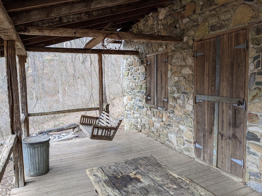 The wooden porch of Myron Glaser cabin shows a wooden, swinging bench and a metal trashcan.