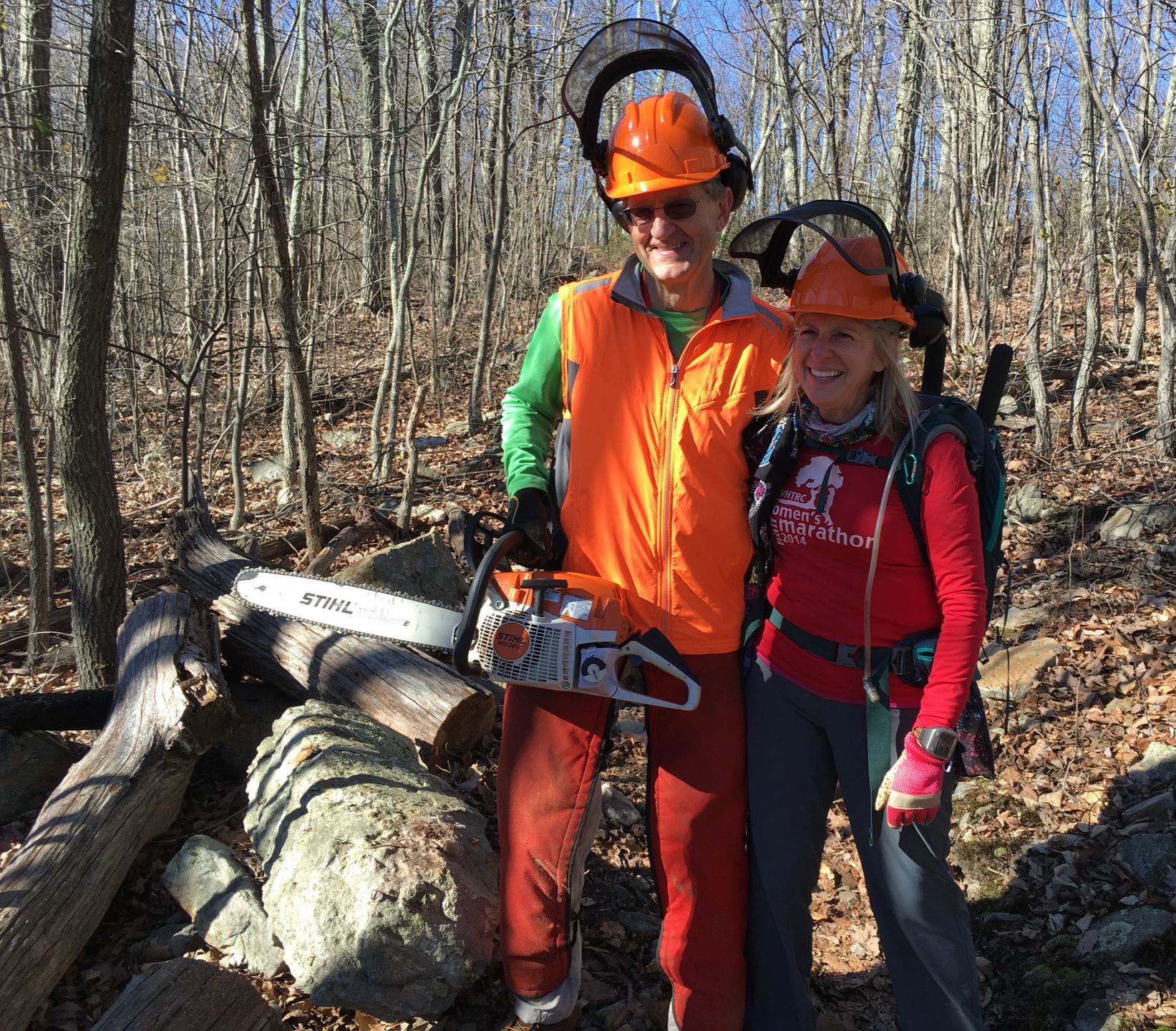 A man and a woman are standing in the woods with a chainsaw.