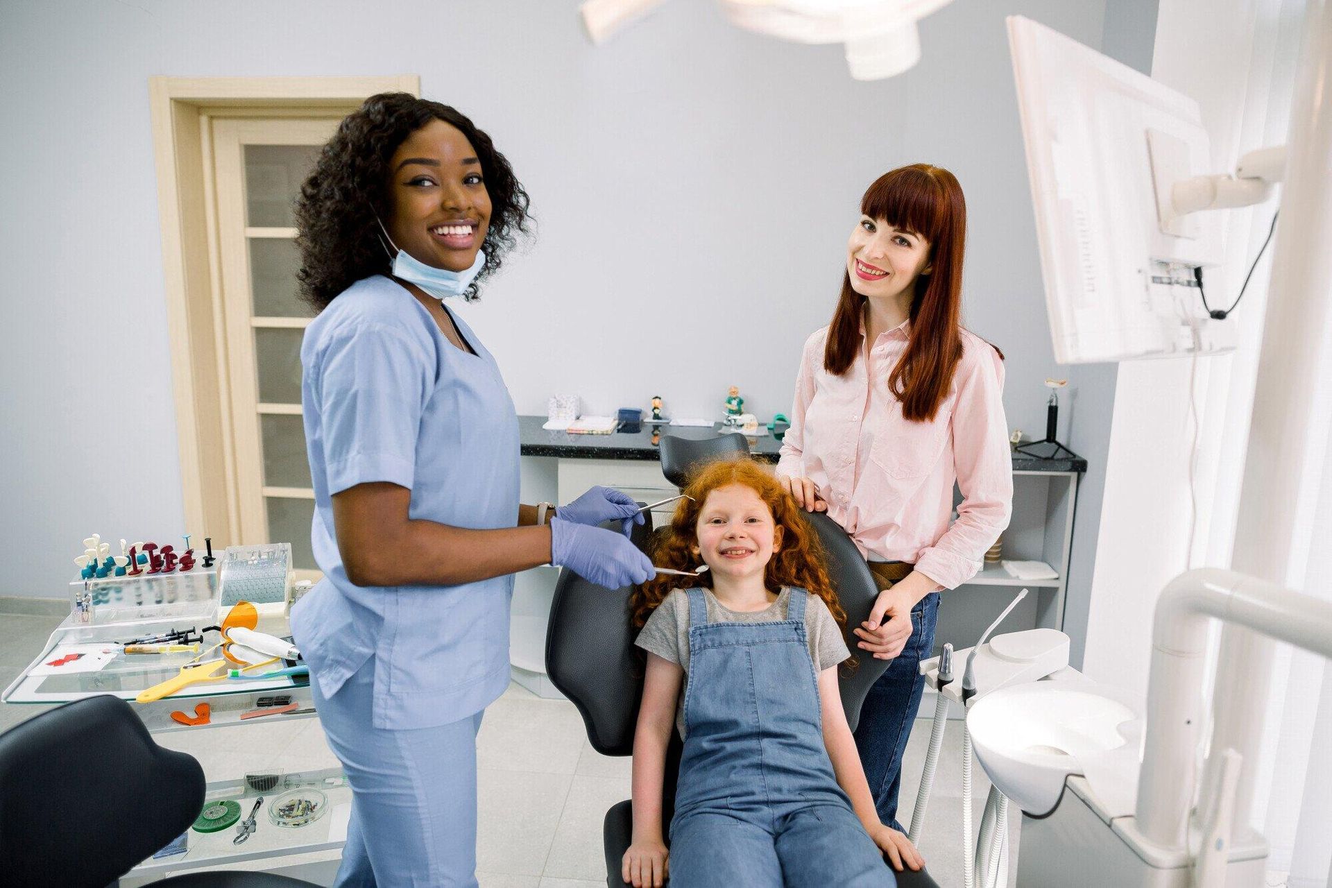 A woman is standing next to a dentist and a little girl in a dental chair.