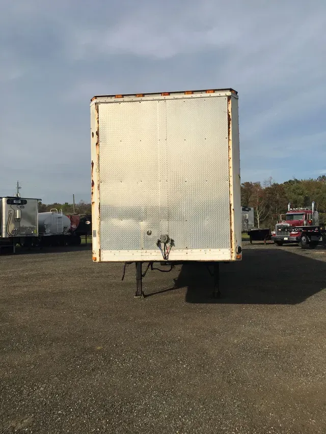 A white, rusty trailer sits on a gravel lot, with a blue sky and other trucks in the background.
