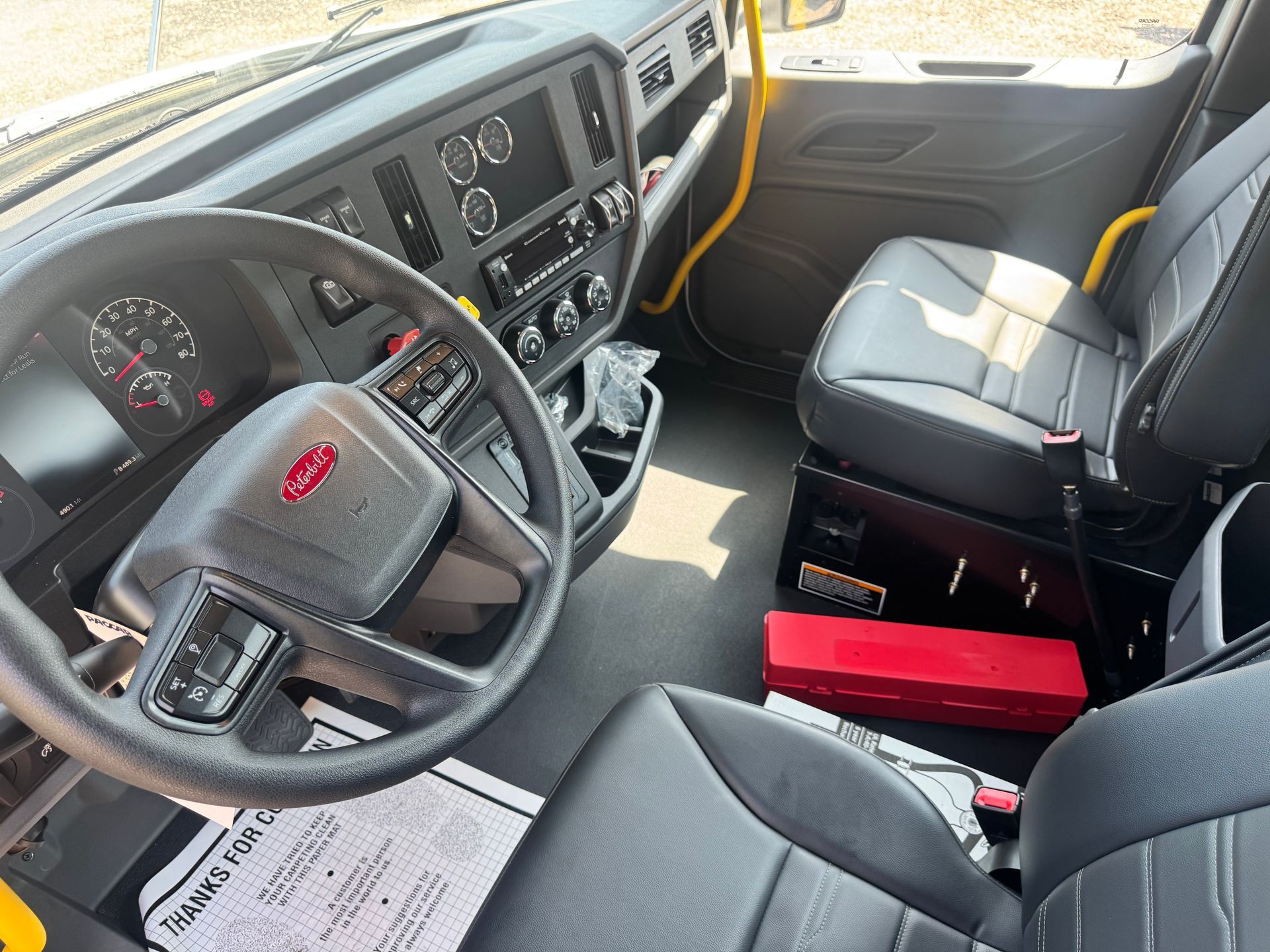 Interior of a Peterbilt truck cab, featuring black seats, dashboard, and steering wheel. A red toolbox is on the floor.