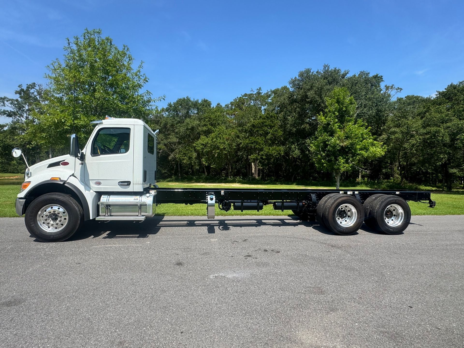 White cab-over truck chassis on asphalt road with a backdrop of green trees and a blue sky.
