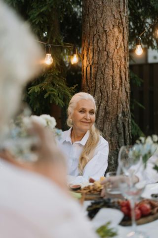 An elderly woman is sitting at a table with a tree in the background.