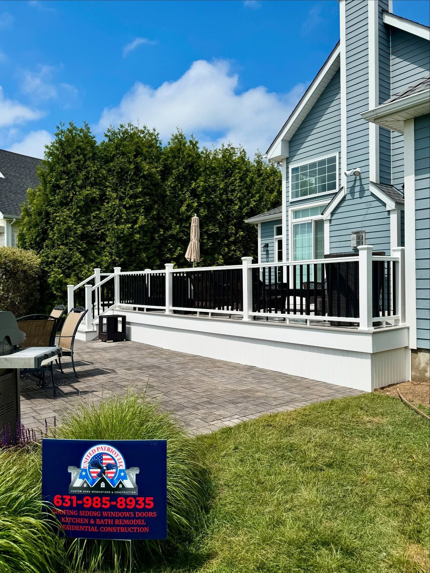 A deck with black railings and white posts attached to a blue house. A sign with a phone number in the foreground.