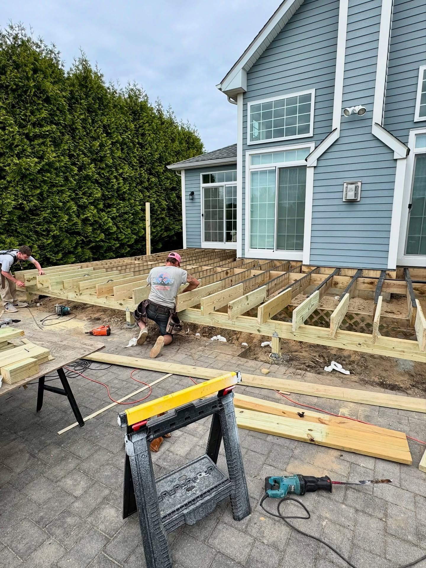 Construction workers building a wooden deck near a blue house. Sawhorses and tools are visible.