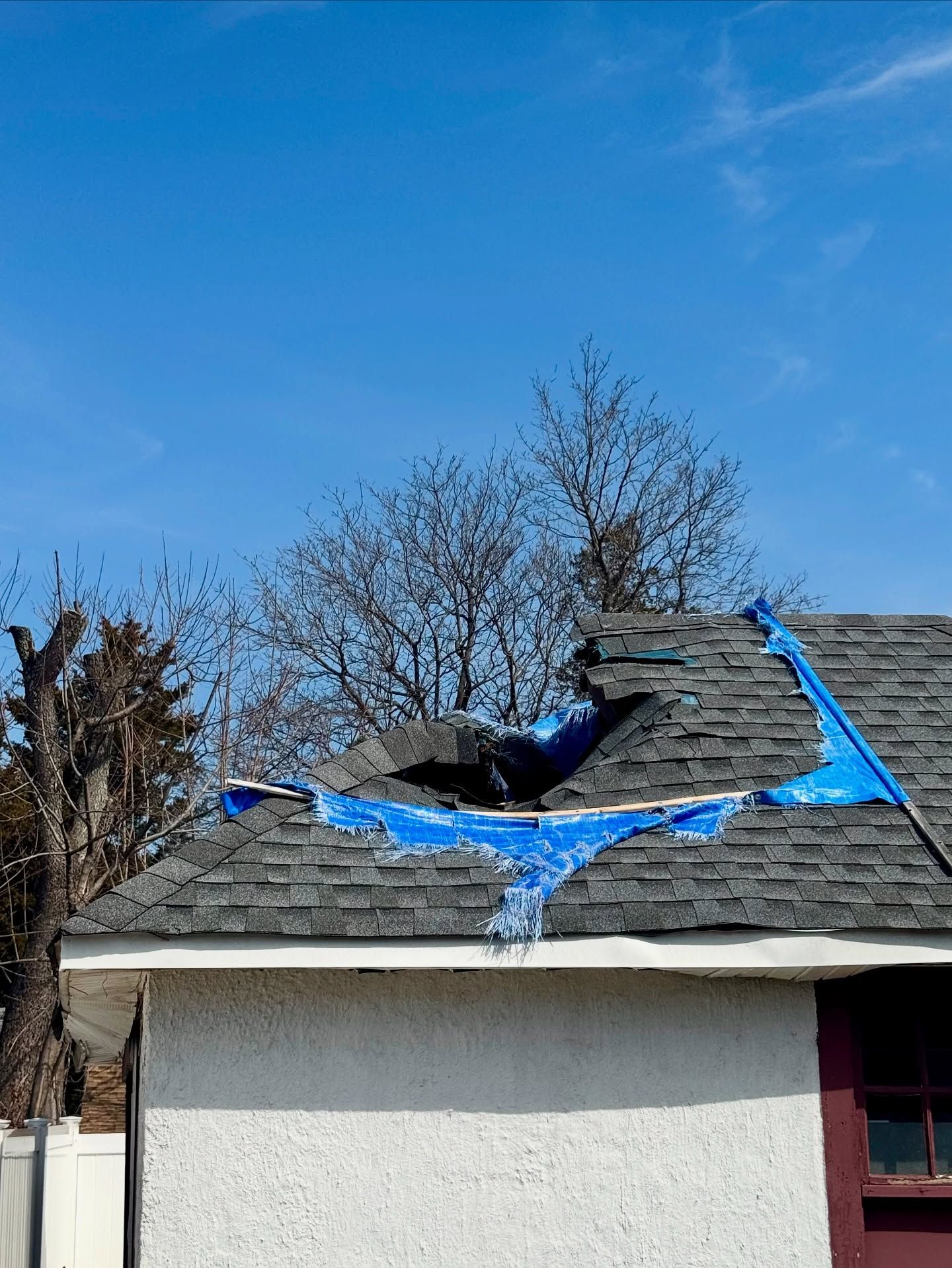 Damaged roof covered by blue tarp against a blue sky, near bare trees.