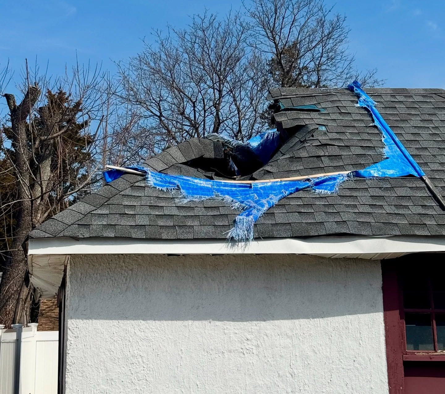 Damaged roof with hole covered by torn blue tarp, against a clear blue sky.