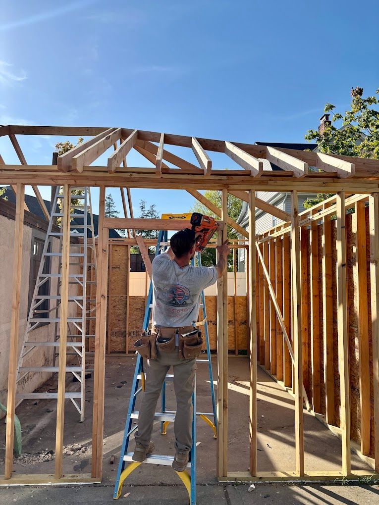 Man on ladder framing a wooden structure with a nail gun on a sunny day.