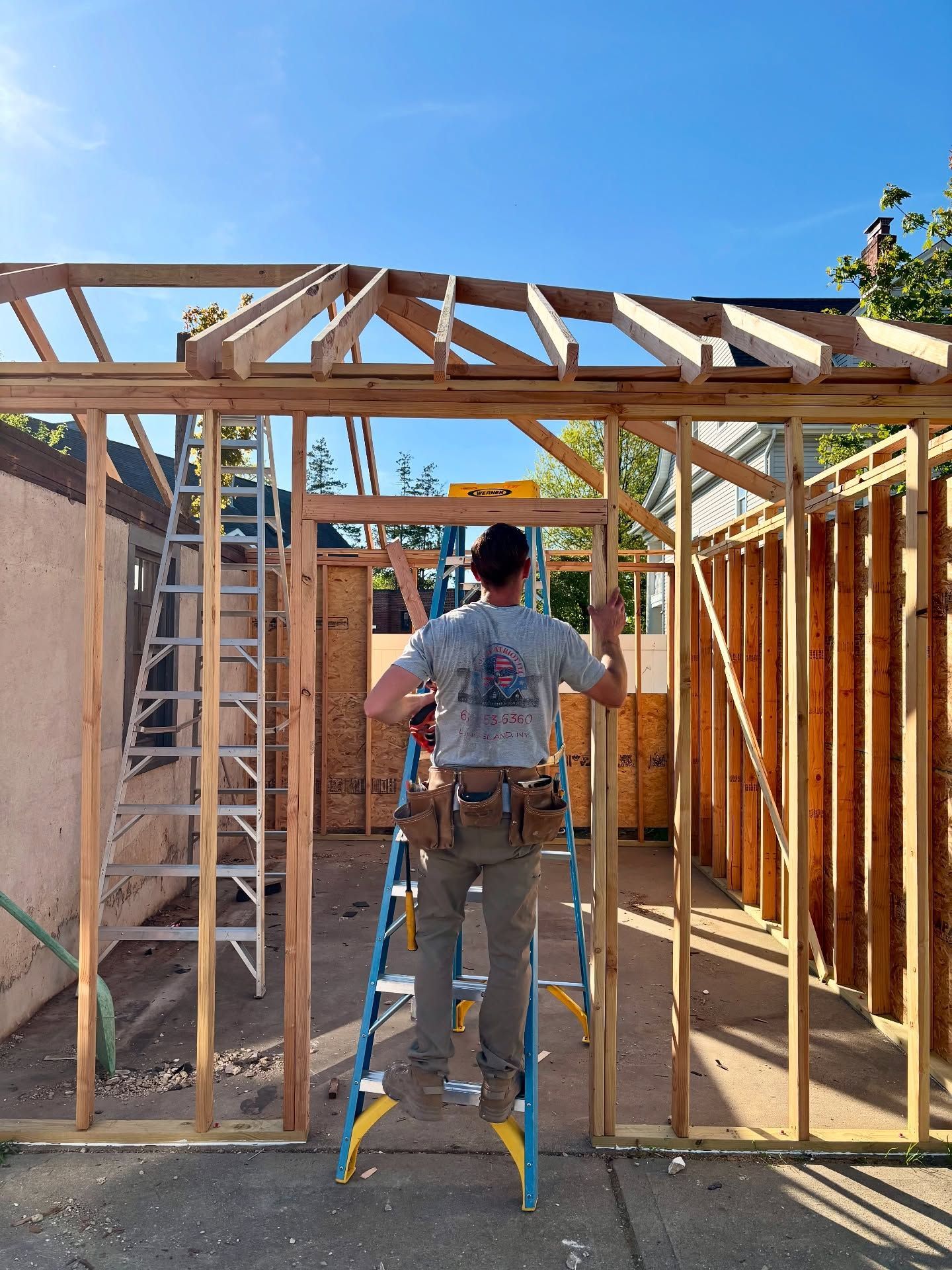 Construction worker framing a structure on a sunny day, standing on a ladder and using tools.
