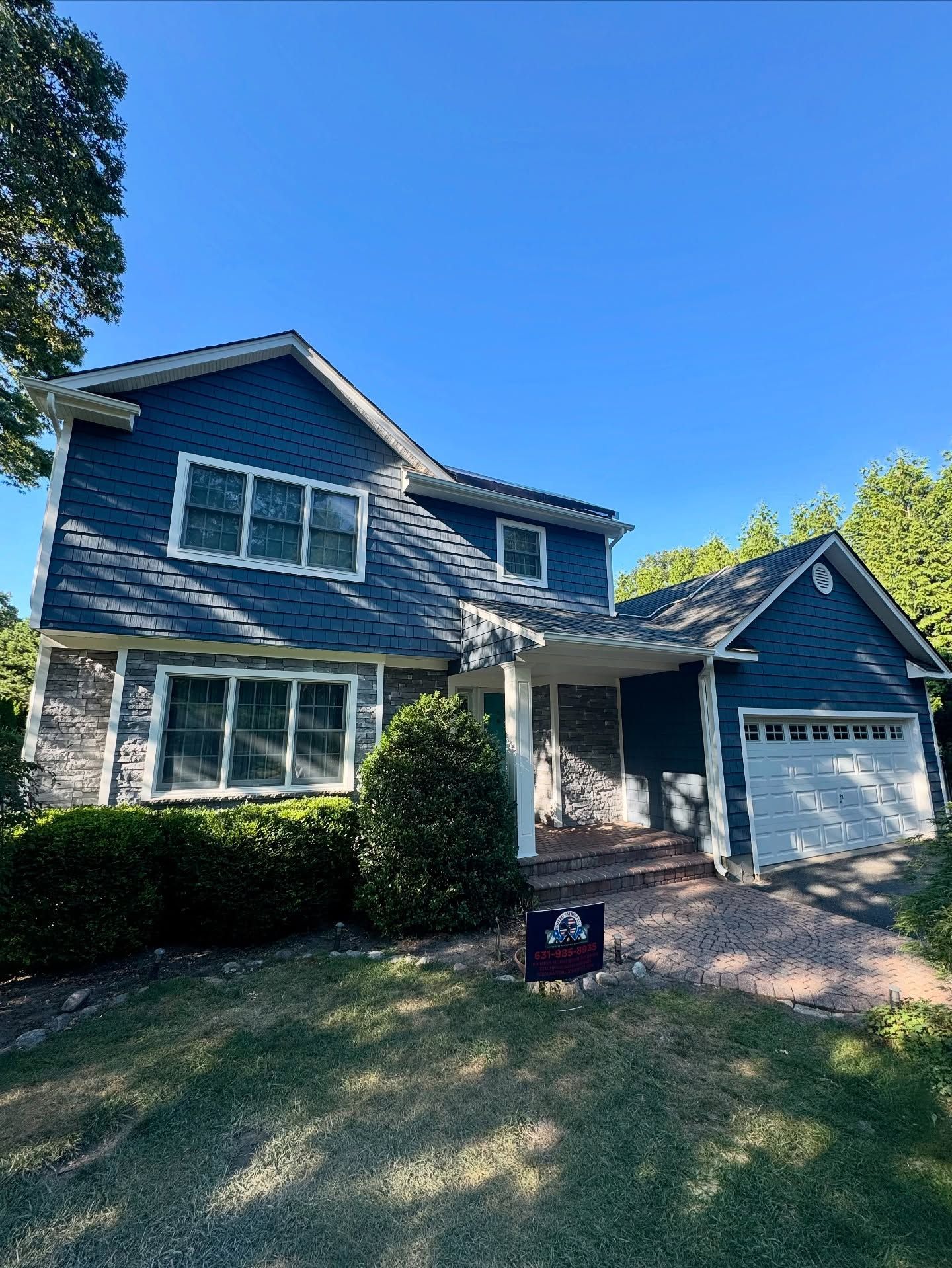 Blue-sided two-story house with a white garage door, surrounded by a lawn and trees under a clear sky.
