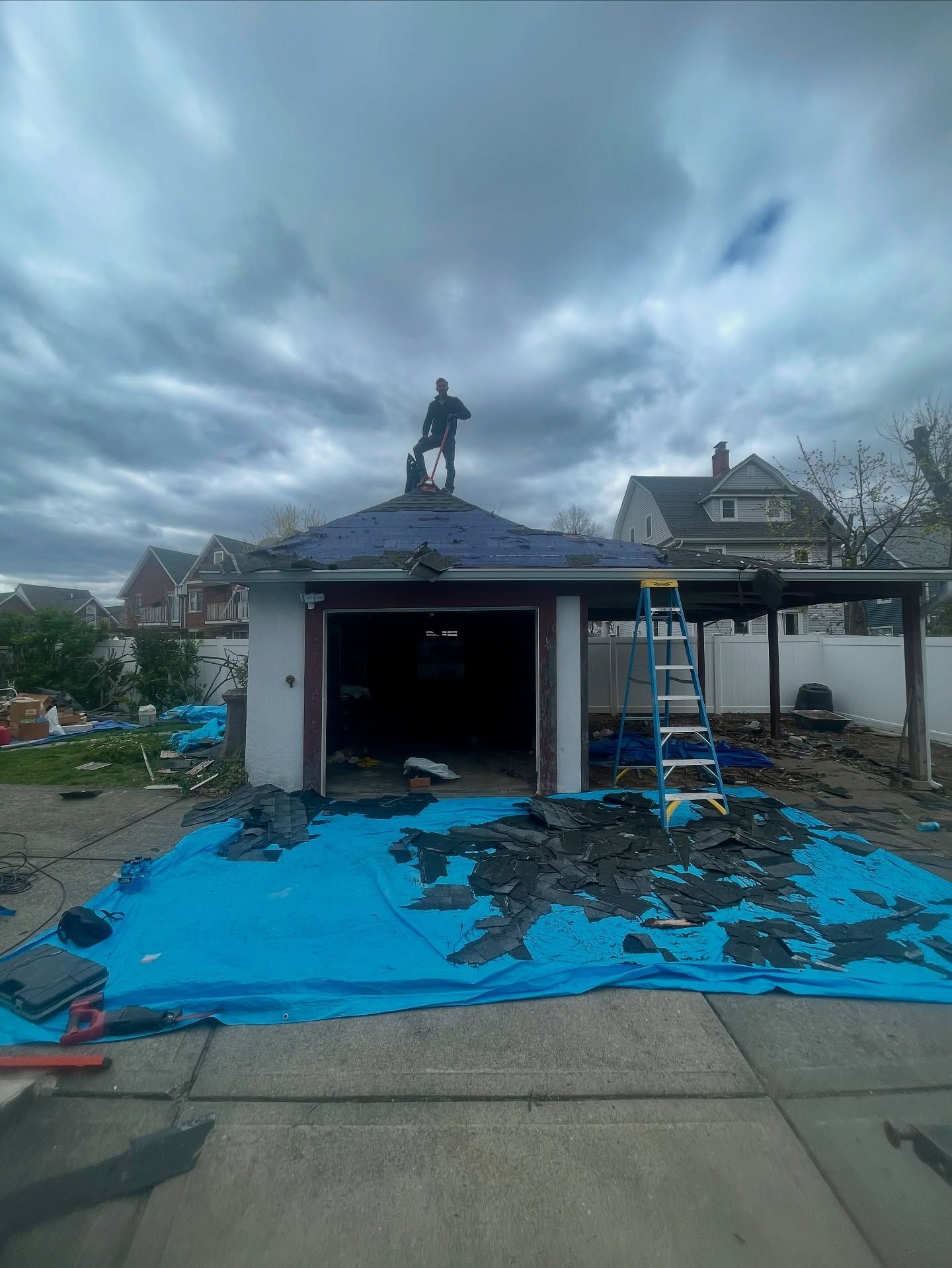 Bryan Tooker stands on a garage roof with a tarp covering debris below. Overcast sky.