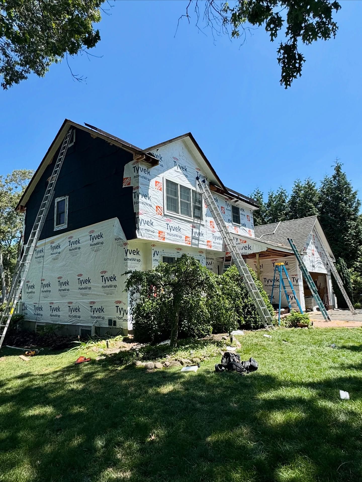 House under construction, covered in white wrap, with ladders against it. Blue siding visible. Sunny day.