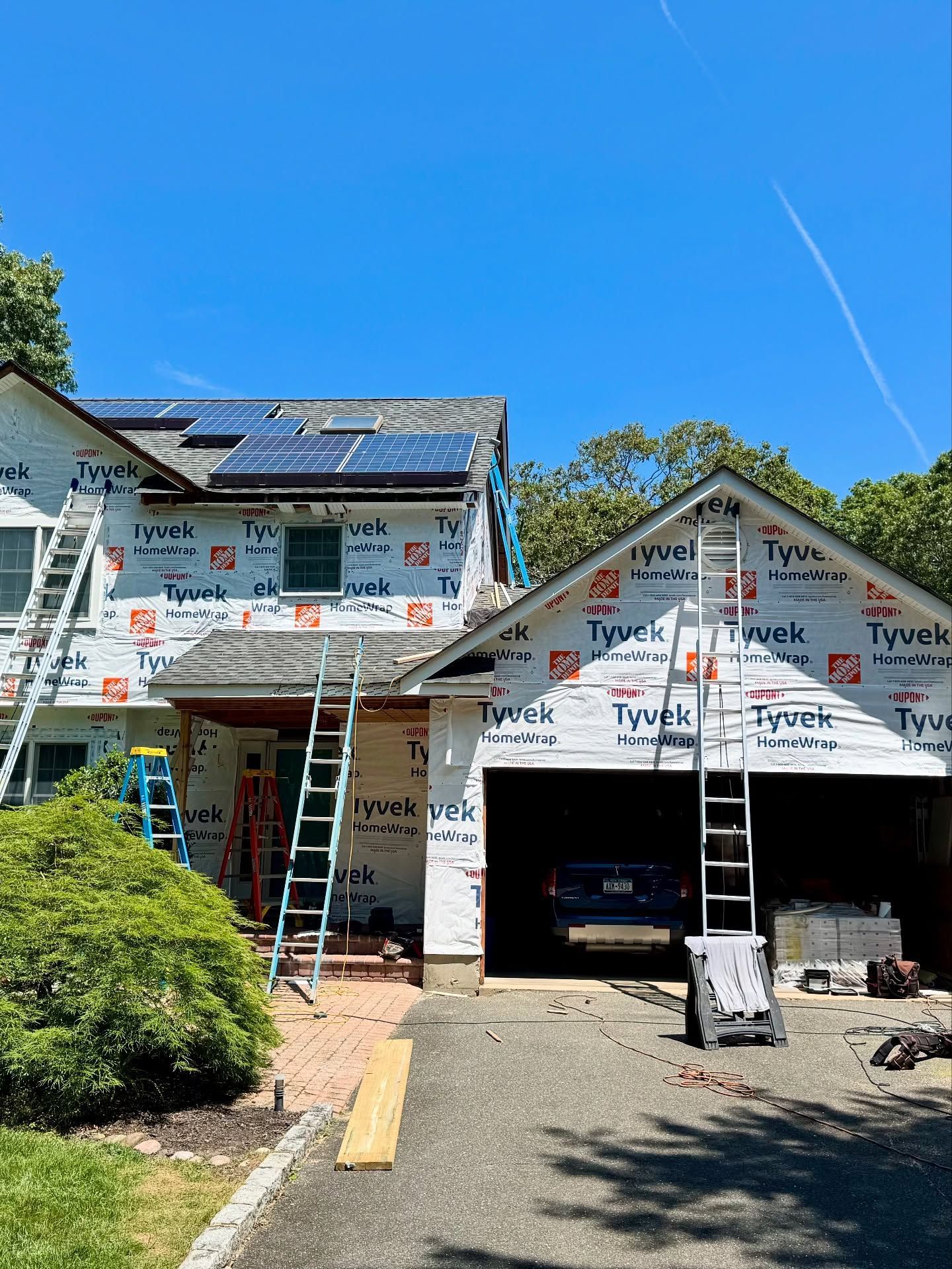 House under construction, covered in Tyvek, with ladders. Solar panels on roof, open garage.