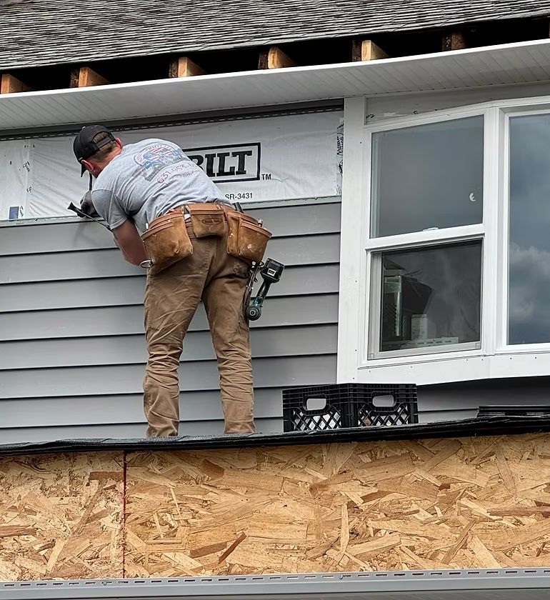Bryan installing siding on a house exterior; gray siding, white window, brown tool belt.