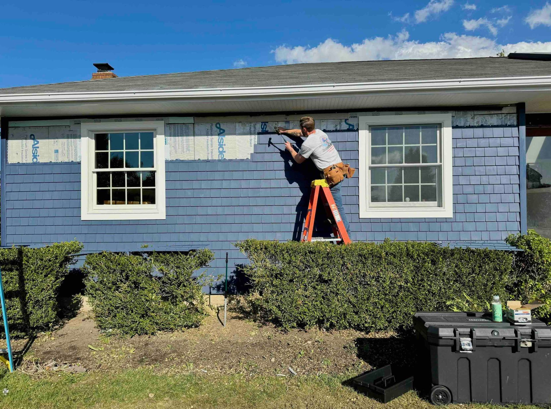 Person on ladder repairing blue shingle siding near two windows. Bright blue sky. Toolbox and bushes in front.