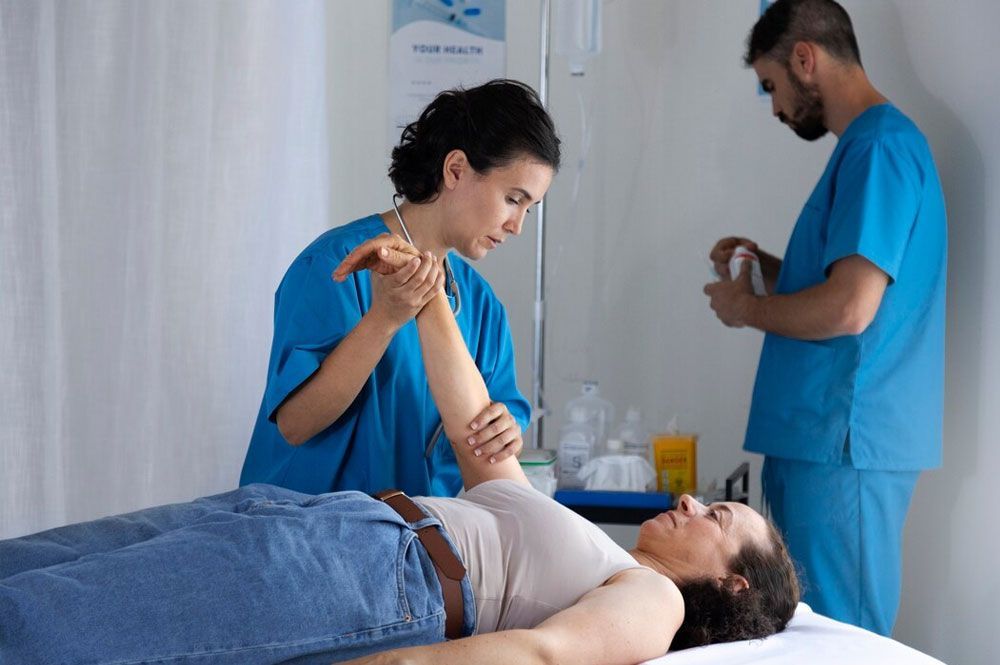 Woman's arm is examined by a medical professional in a hospital setting; another person prepares something nearby.