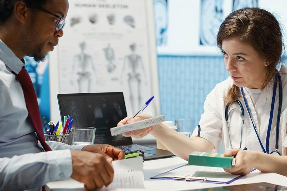 Doctor hands a patient a medication box. Medical office setting with a skeletal chart visible.