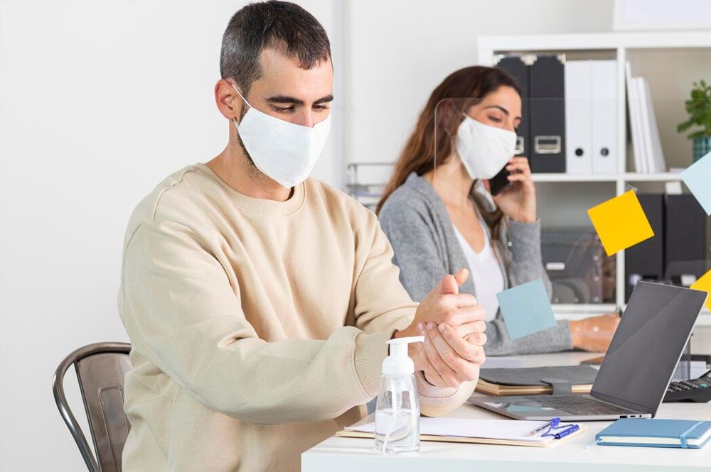 Man applying hand sanitizer at a desk, masked coworker on the phone nearby. Office setting.