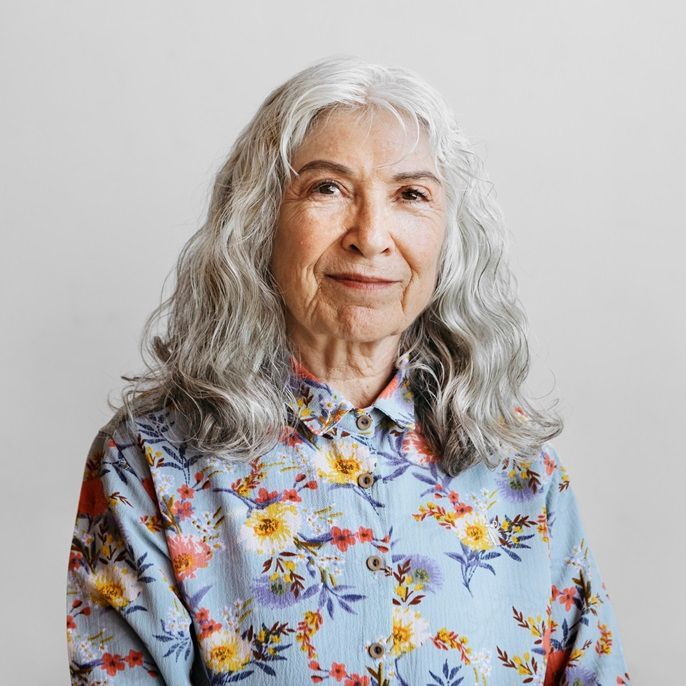 Woman with long gray hair smiles, wearing a blue floral shirt, against a light gray backdrop.