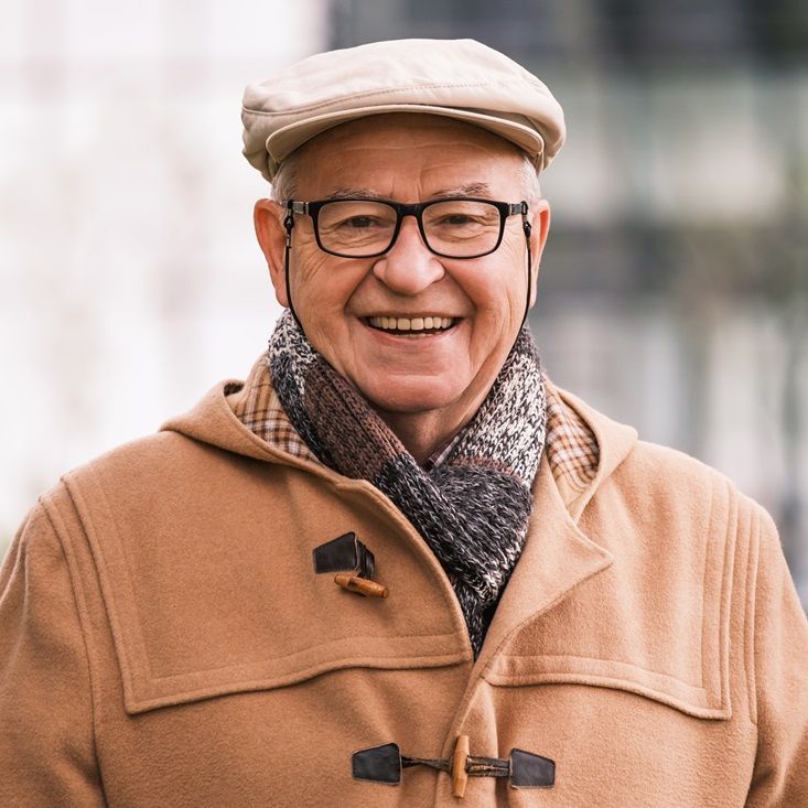 Smiling man wearing glasses, beige cap, coat, and scarf; outdoors.