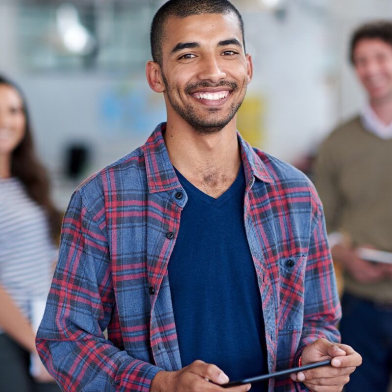 Man smiling, holding a tablet, wearing a plaid shirt and blue top. Other people in the background.