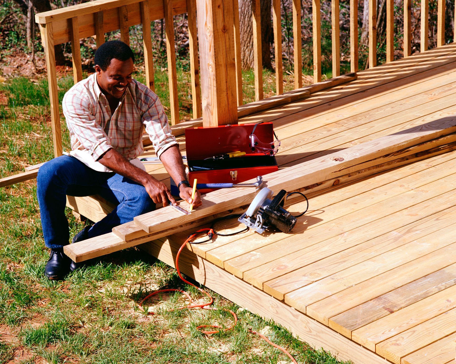 A man sits on a wooden deck working on a piece of wood