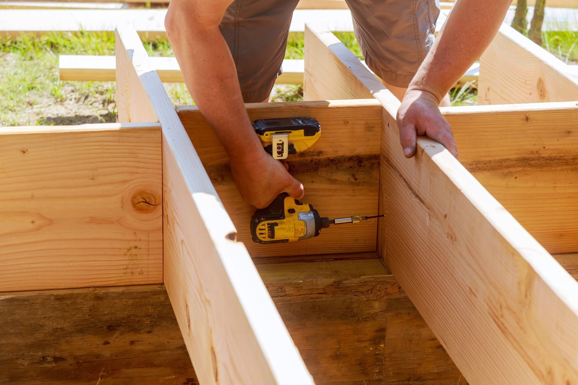 A male worker using a yellow drill to assemble wooden beams outdoors.