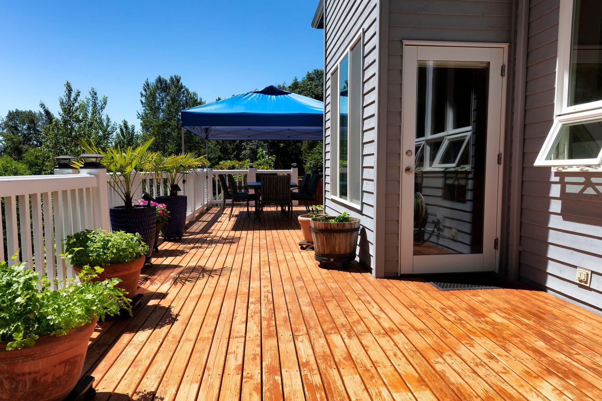 Wooden deck with potted plants, table, chairs, and a blue umbrella on a sunny day.
