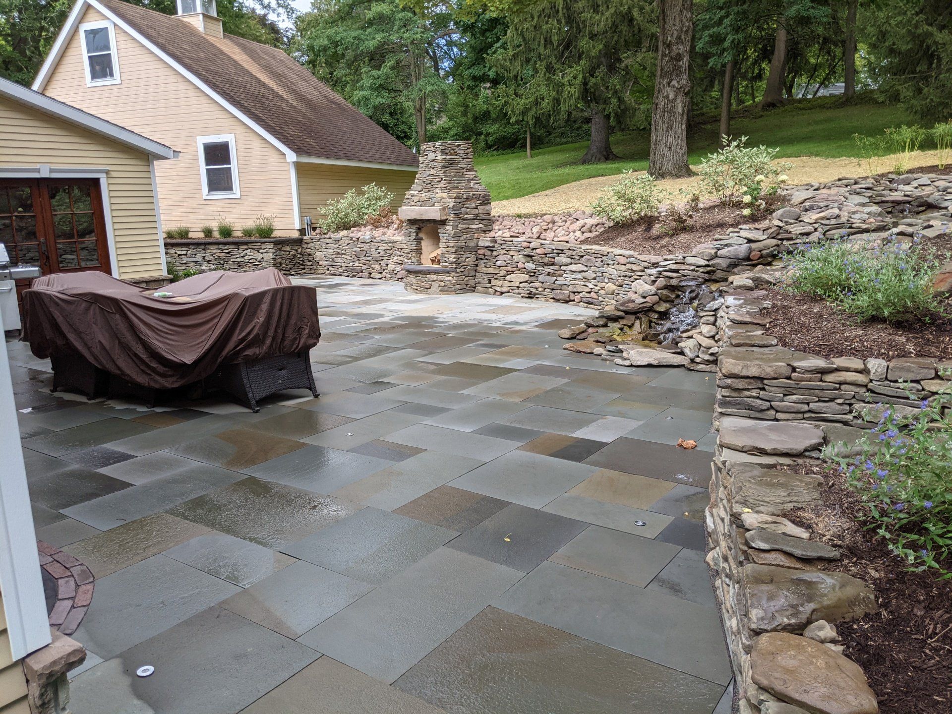 a natural stone patio with a table and chairs and a house in the background and a retaining wall 