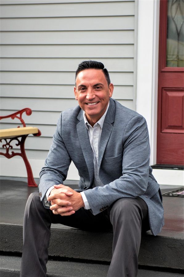 Man sitting on front steps in a gray blazer, smiling beside a red door and white siding.