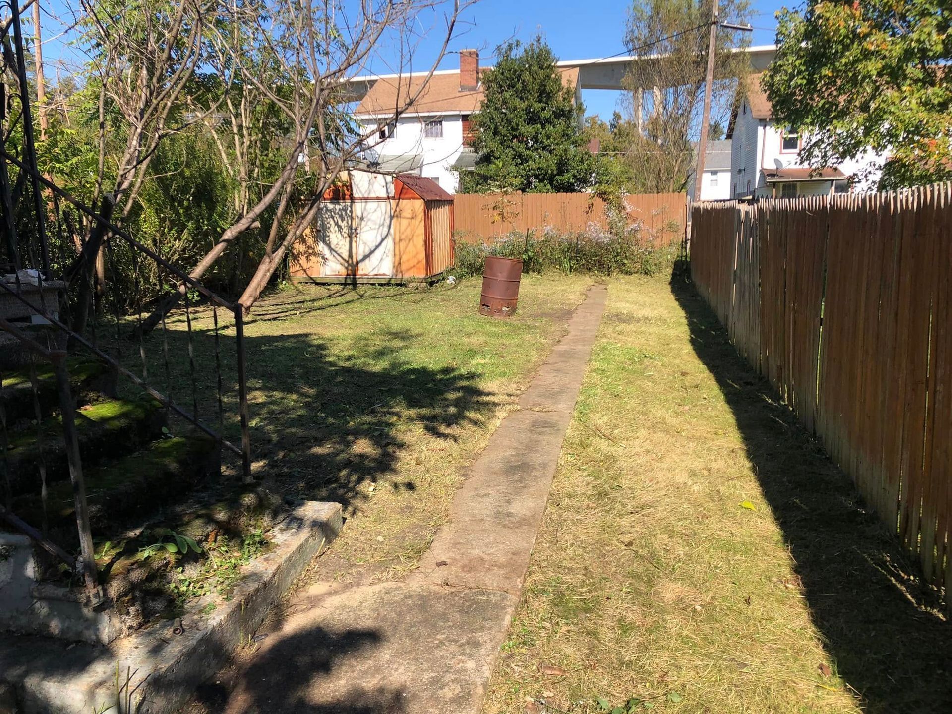 A backyard path leads past a small shed toward a wooden fence, with houses visible in the distance under a blue sky.