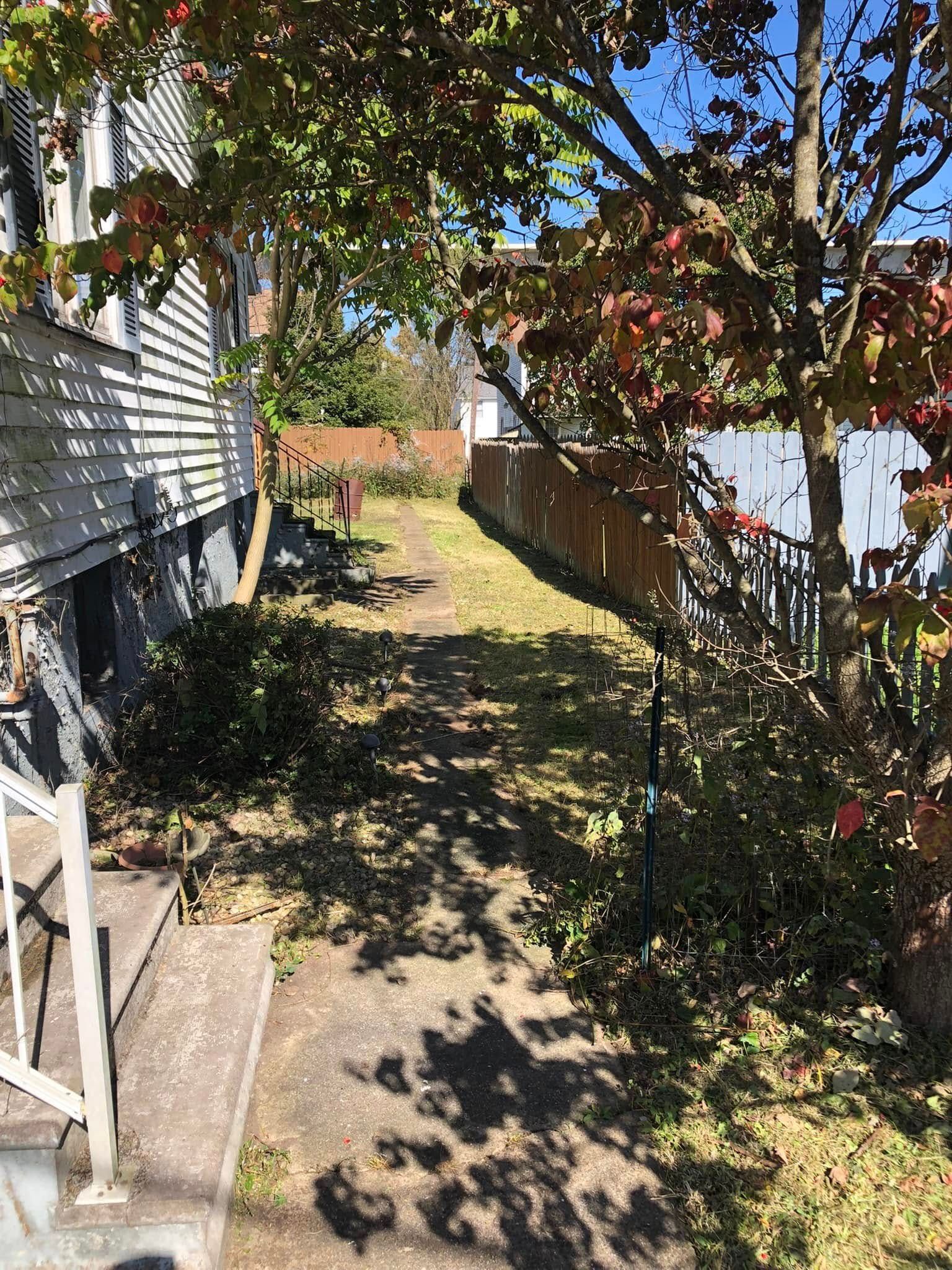 A narrow dirt path runs between the side of a white house with stairs and a wooden fence, shaded by a tree with red leaves.
