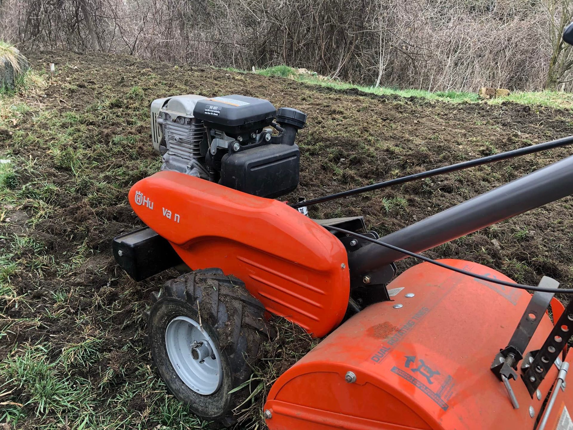 A bright orange walk-behind rotary tiller sits in a freshly plowed field of dark soil.