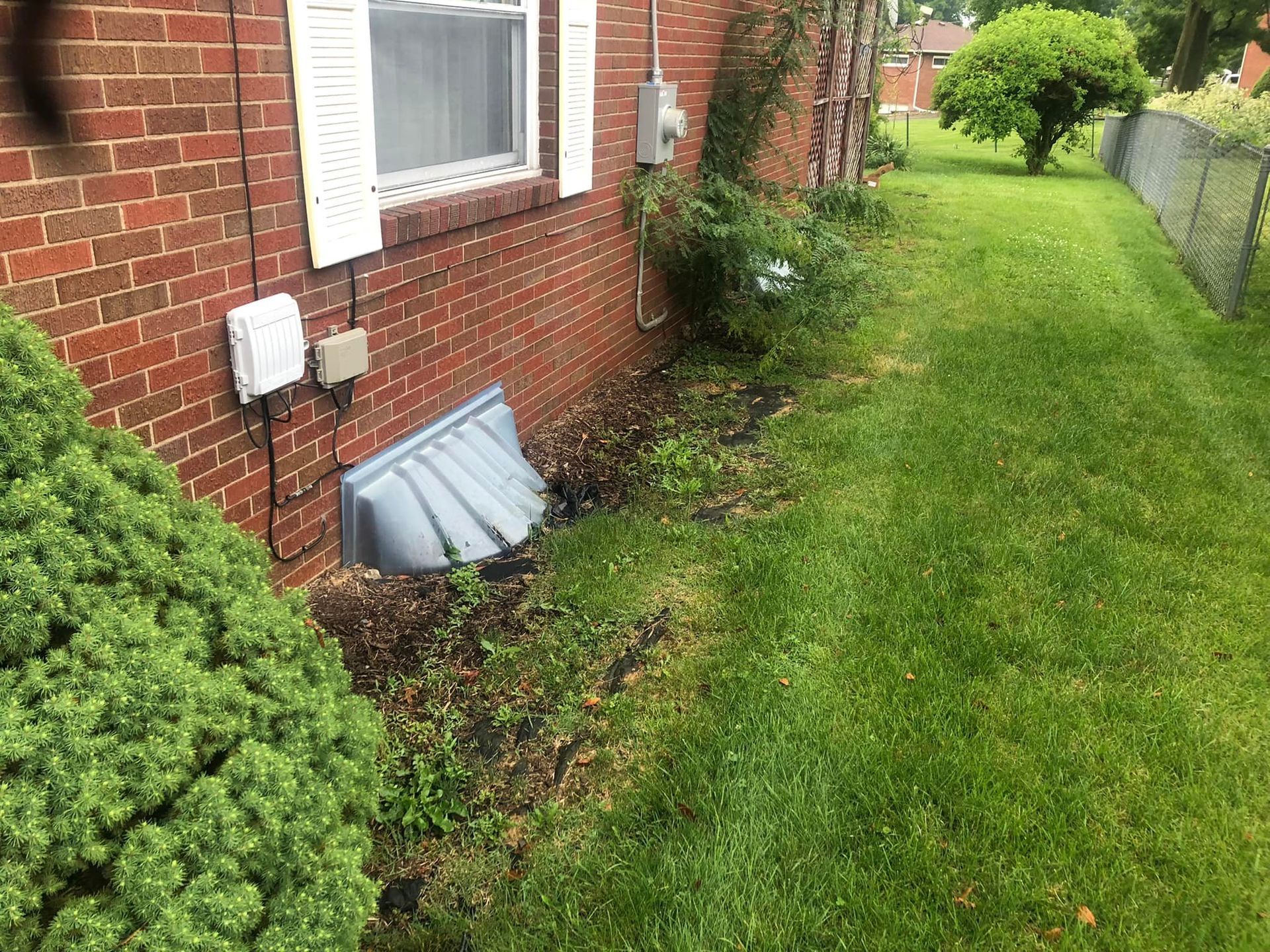 A red brick house exterior with a gray plastic window well cover, electrical utility boxes, and a green grass lawn.