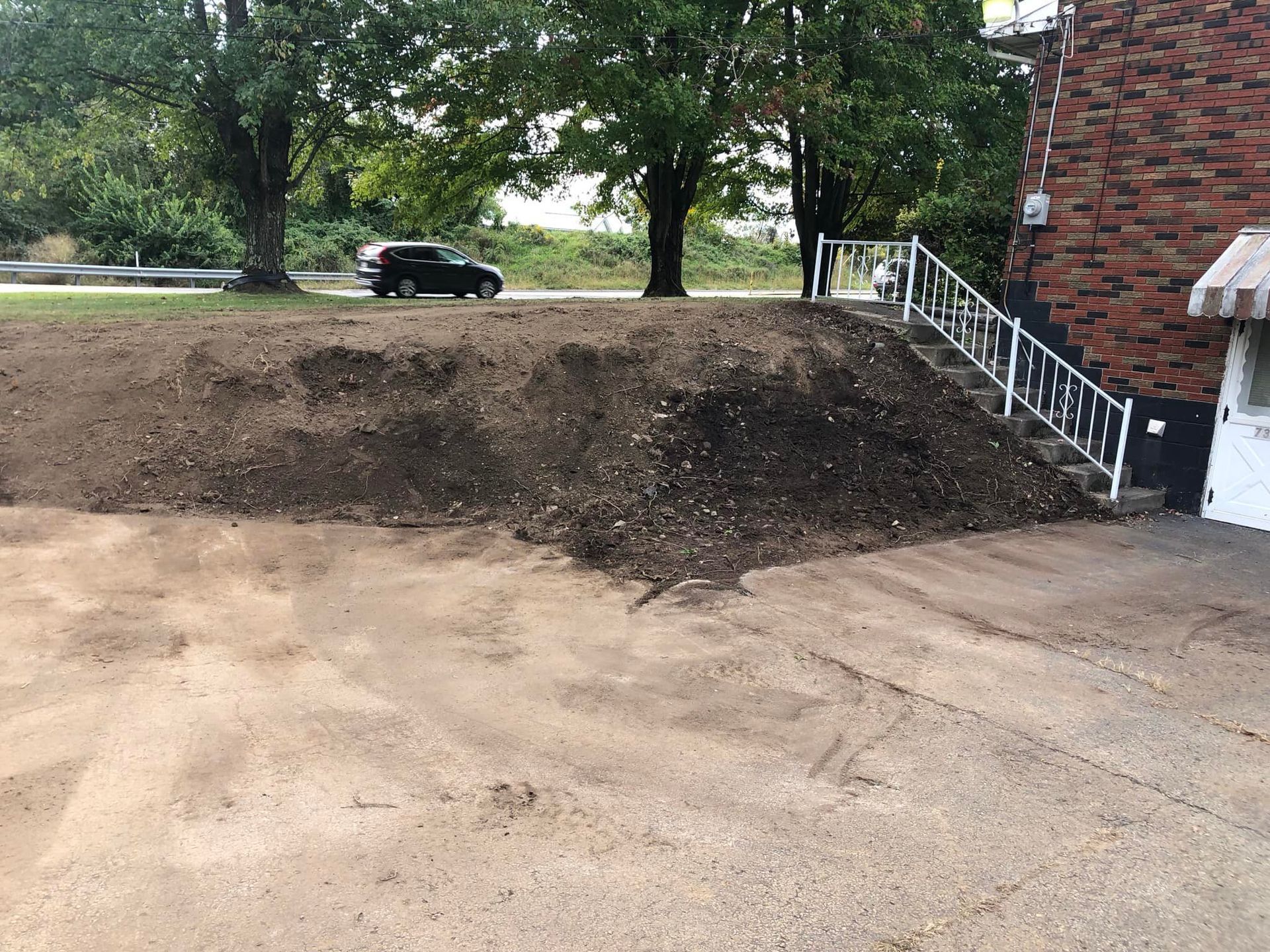 A mound of soil sits next to a brick building with white railings and stairs, with trees and a parked car in the back.