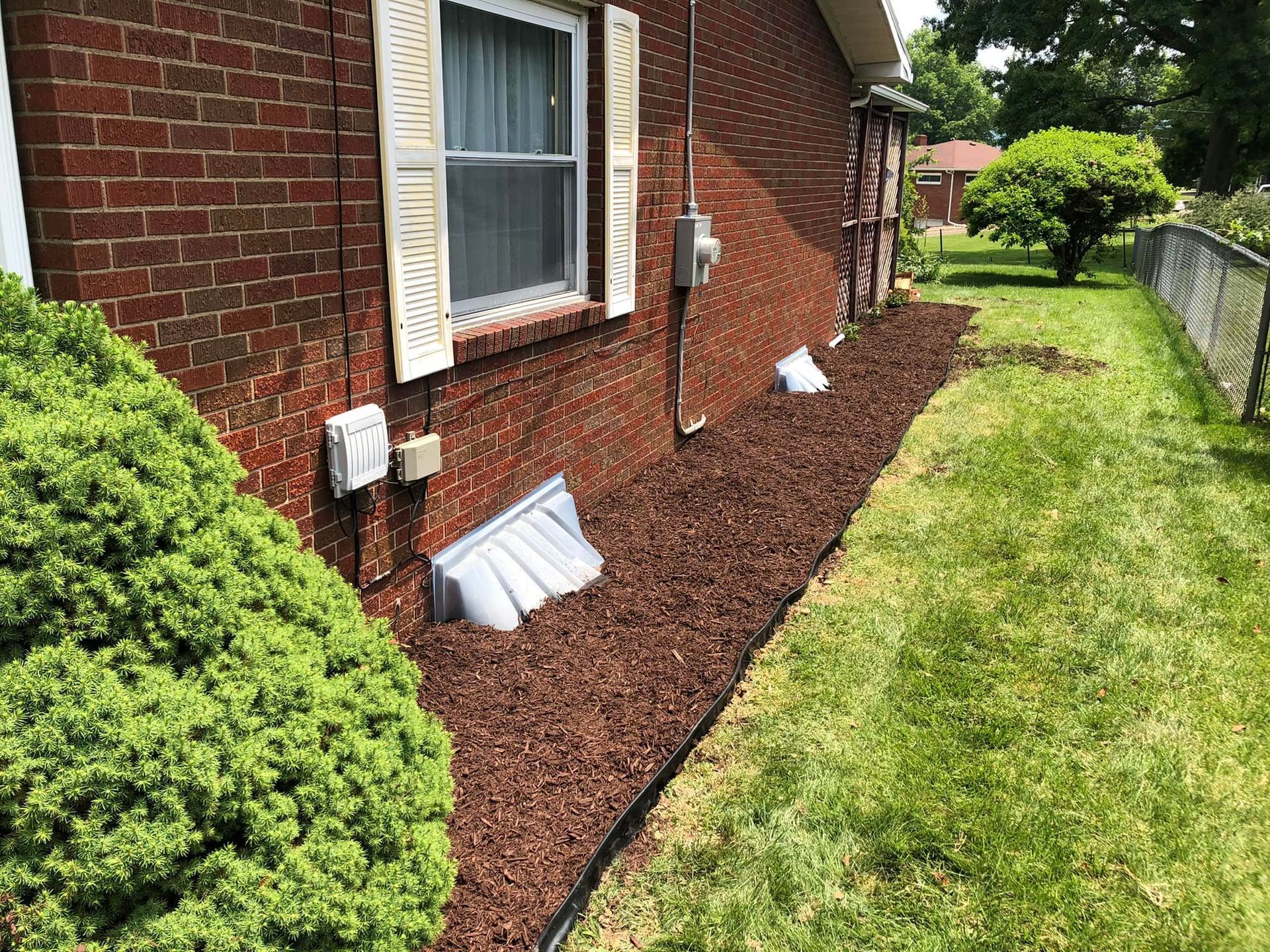 A side view of a brick house featuring a window with shutters, two basement window wells, and a fresh layer of mulch.