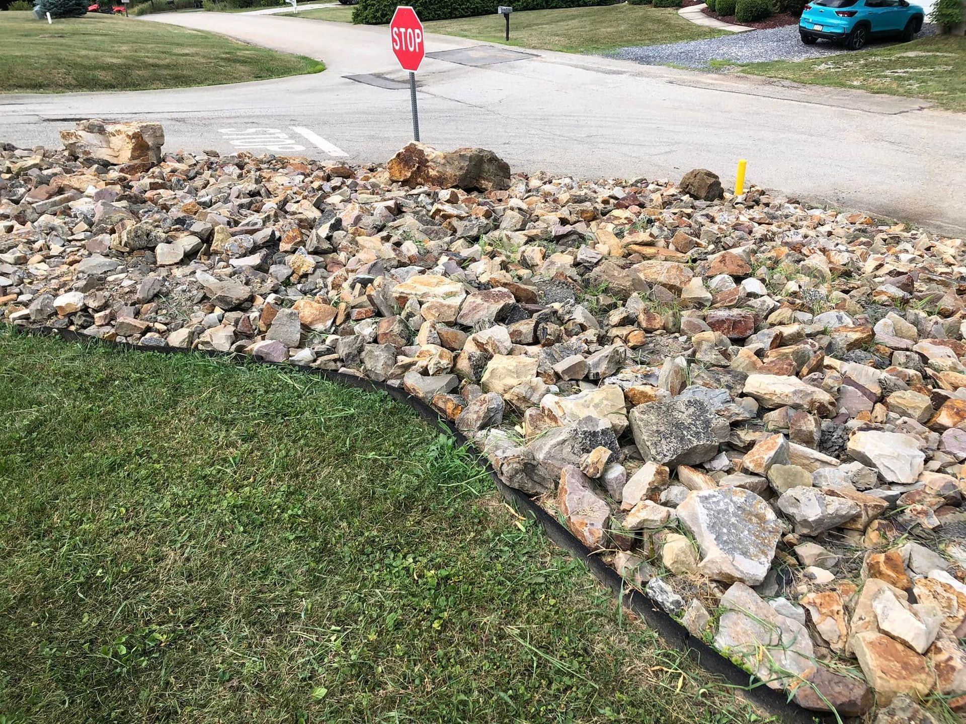 A bed of multi-colored landscaping rocks borders a lawn next to a residential street with a stop sign in the background.