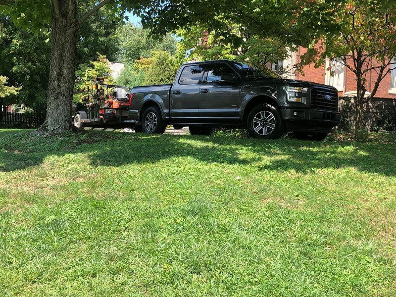 A dark gray pickup truck parked on a grassy lawn with a trailer attached, shaded by large trees.