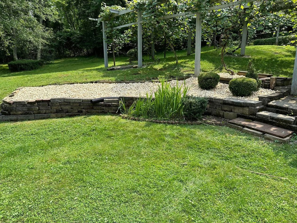 A raised garden bed with stone walls and gravel, featuring small green shrubs and stairs leading up to a grassy lawn.